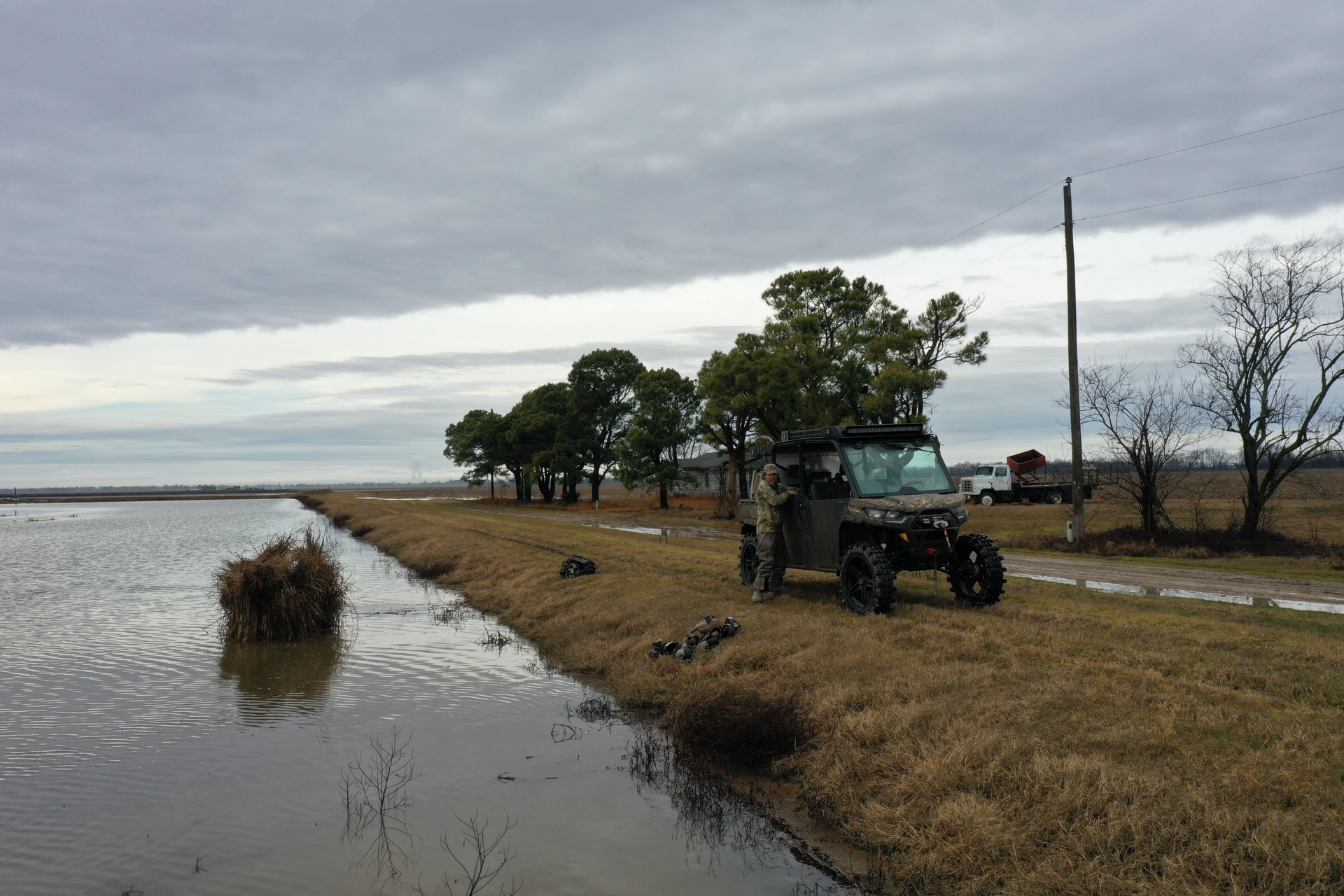 Flooded roadside beside a grassy levee, with a tractor on a dirt road under a cloudy sky.