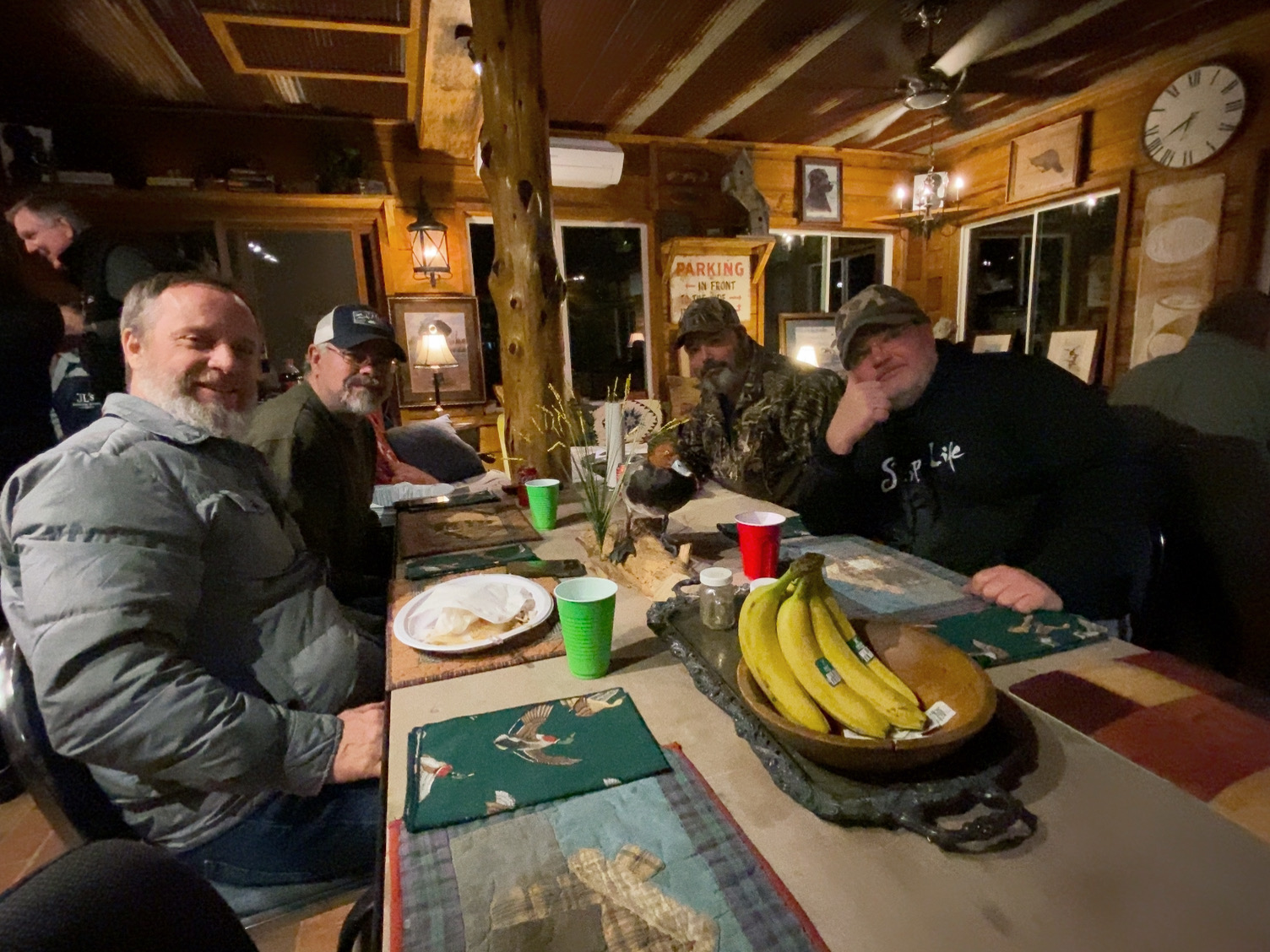 Four people seated around a rustic table in a cozy wooden cabin, with bananas and drinks in the foreground.