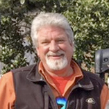 Smiling white-haired man in a brown jacket and orange shirt outdoors