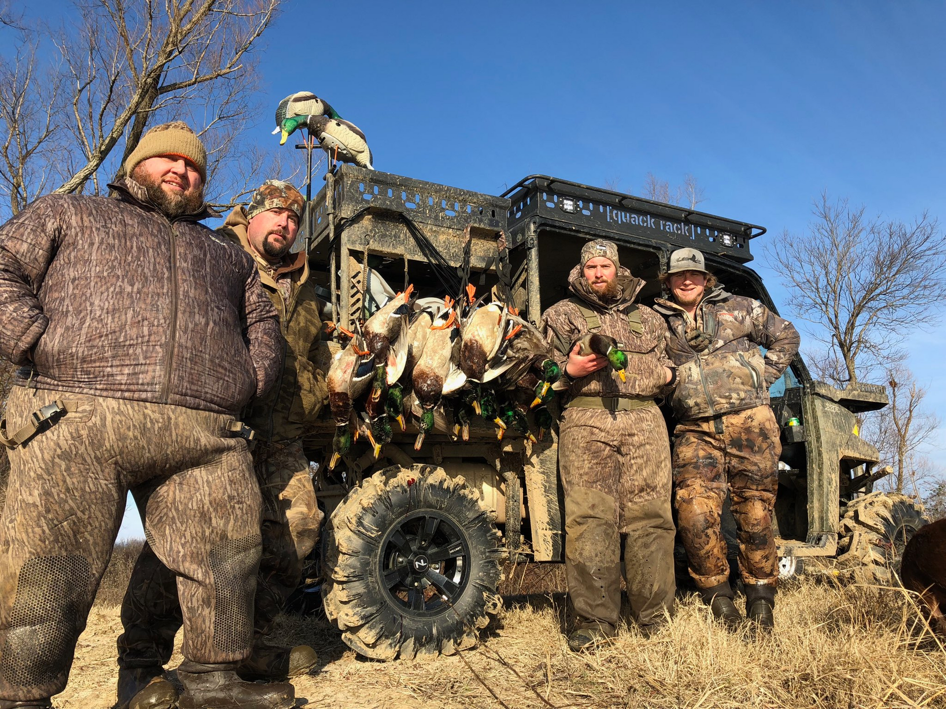Five hunters posing with bird dogs beside an off-road vehicle in a muddy field under a blue sky