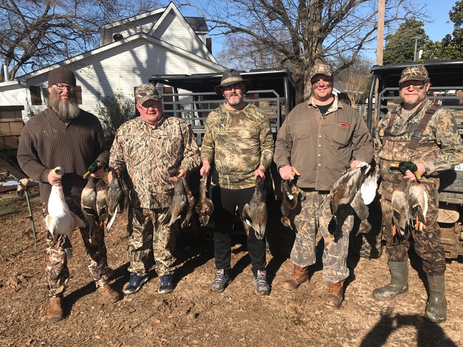 Five hunters in camouflage pose outdoors, holding harvested ducks in a yard near a house.