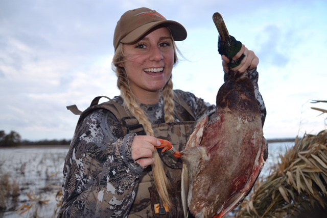 Smiling hunter in camouflage holding a duck by a marsh at dusk