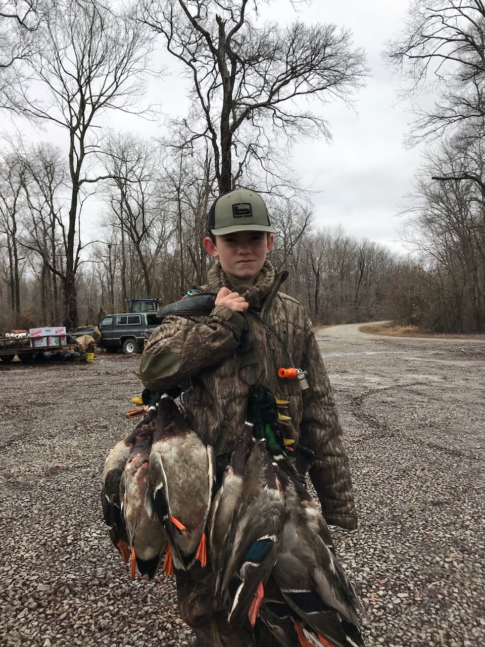 Person holding a large bundle of harvested ducks in a wooded outdoor area, wearing camouflage and a green cap
