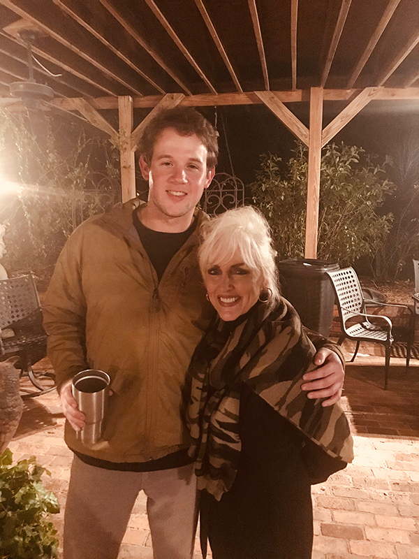 Two people smiling on a brick patio at night, posing under a wooden pergola, one holding a metal cup