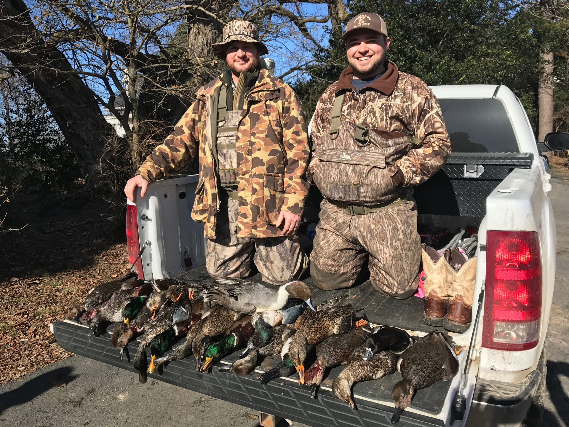 Two hunters pose by a pickup truck bed full of ducks in a wooded area.