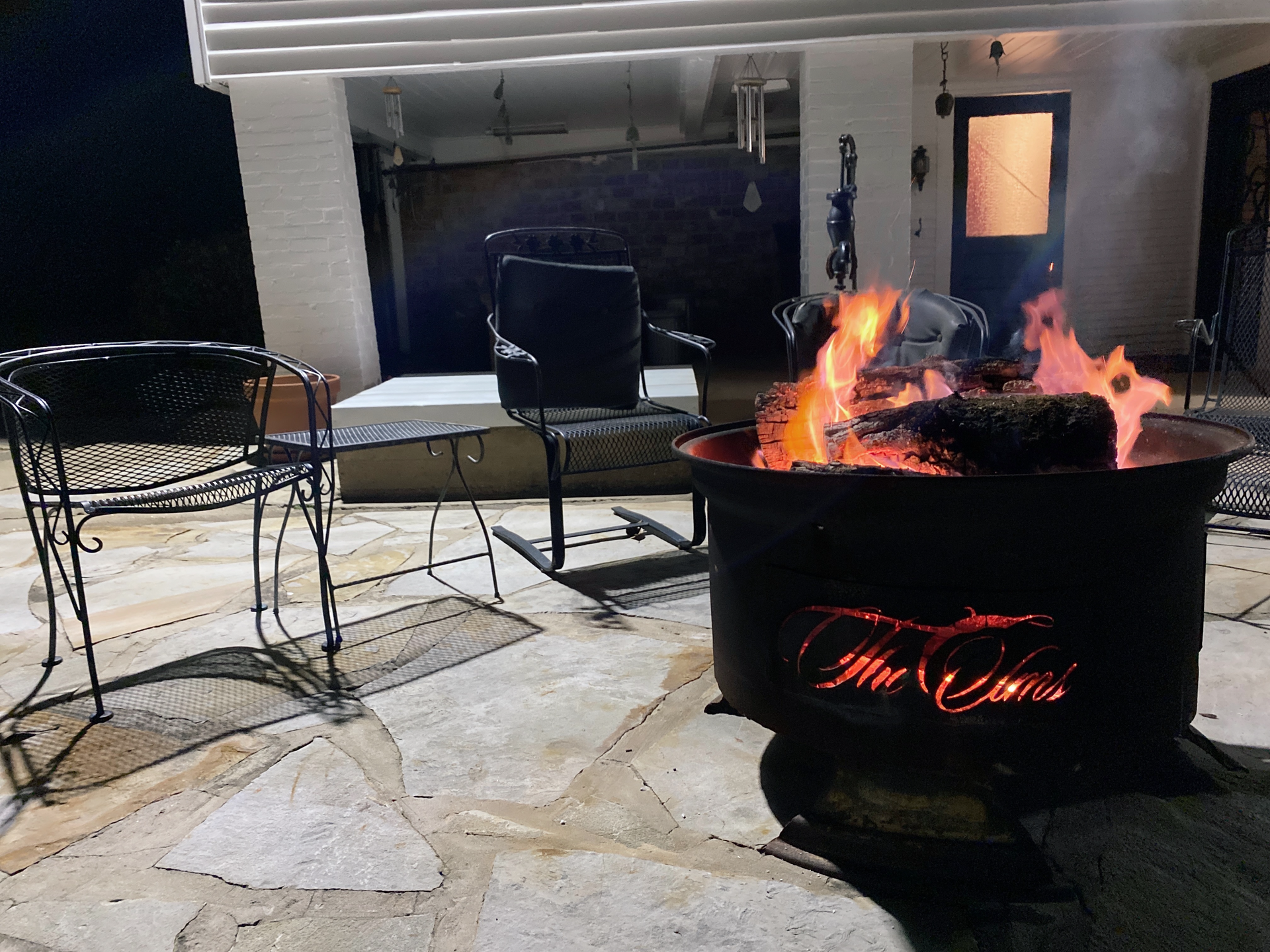 Outdoor patio with a lit fire pit, black chairs, and a table beside a house at night