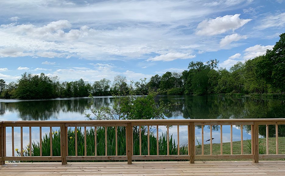 Wooden railing overlooking a calm lake with trees and cloudy blue sky in the background