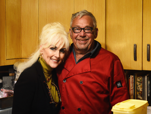 Two smiling people standing together in a kitchen, one in a red shirt and one in black with blonde hair