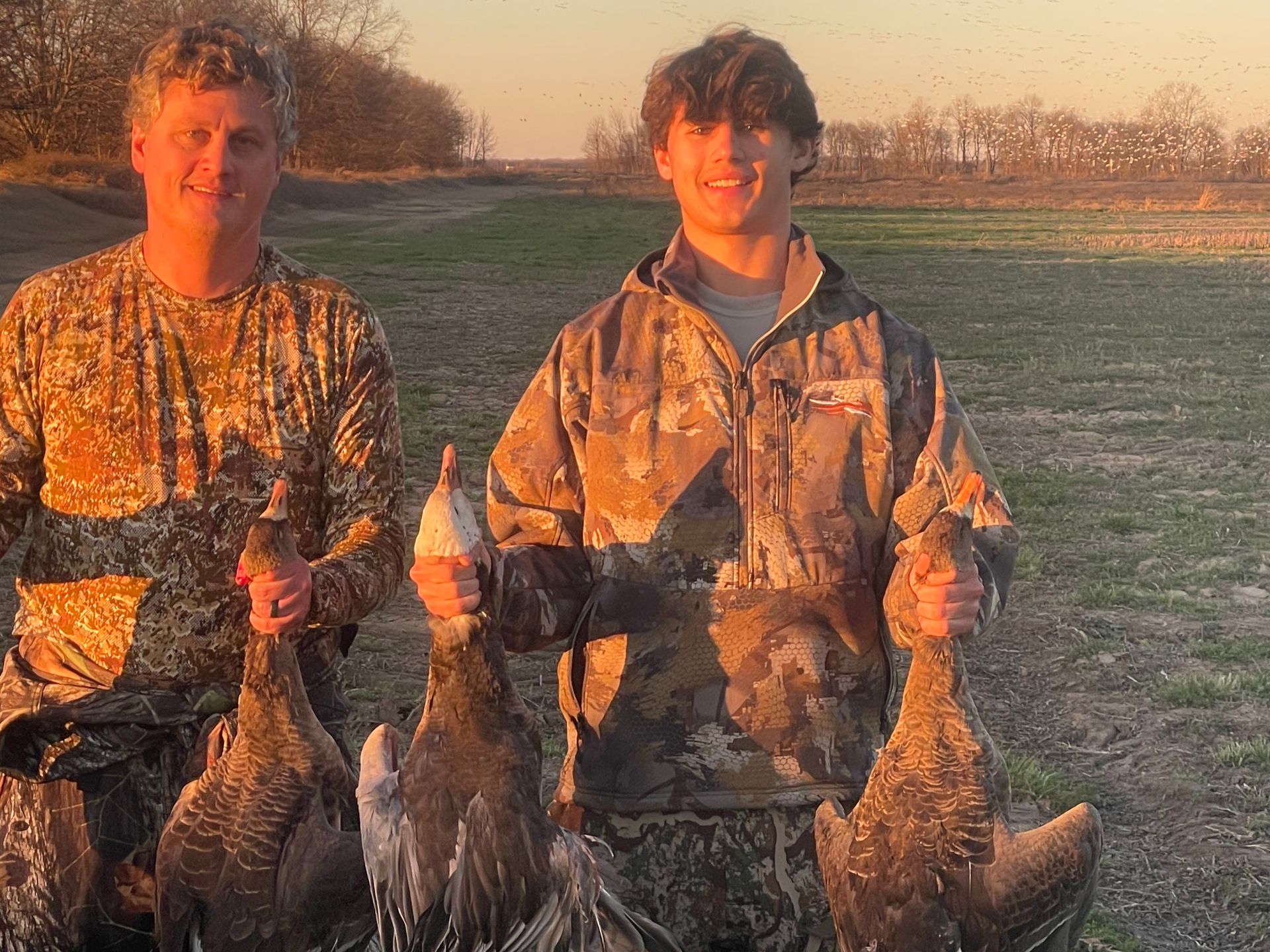Two hunters in camouflage hold several pheasants at sunset in a field.