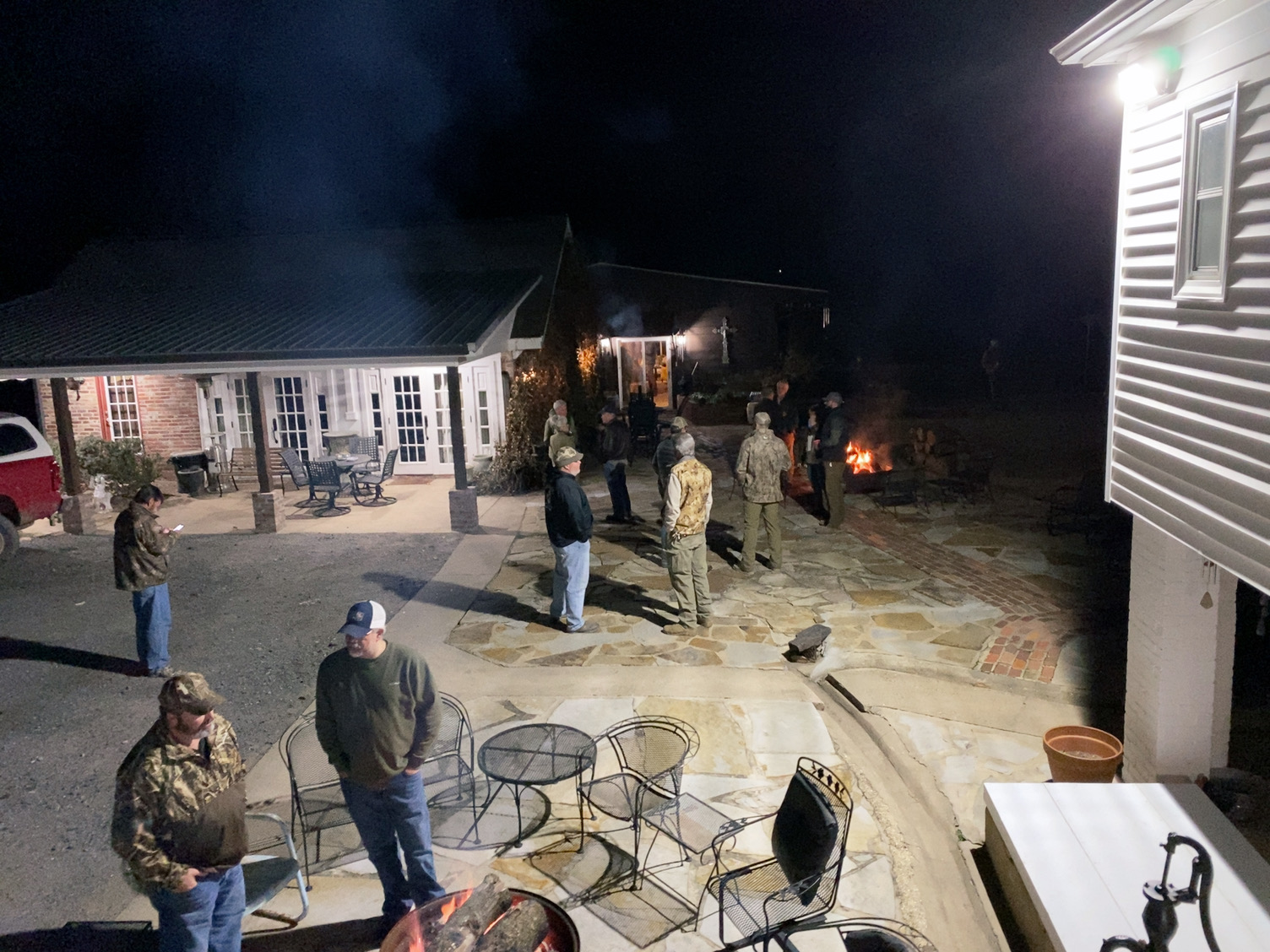 People gathered outside at night around patio tables near a lit house and driveway.