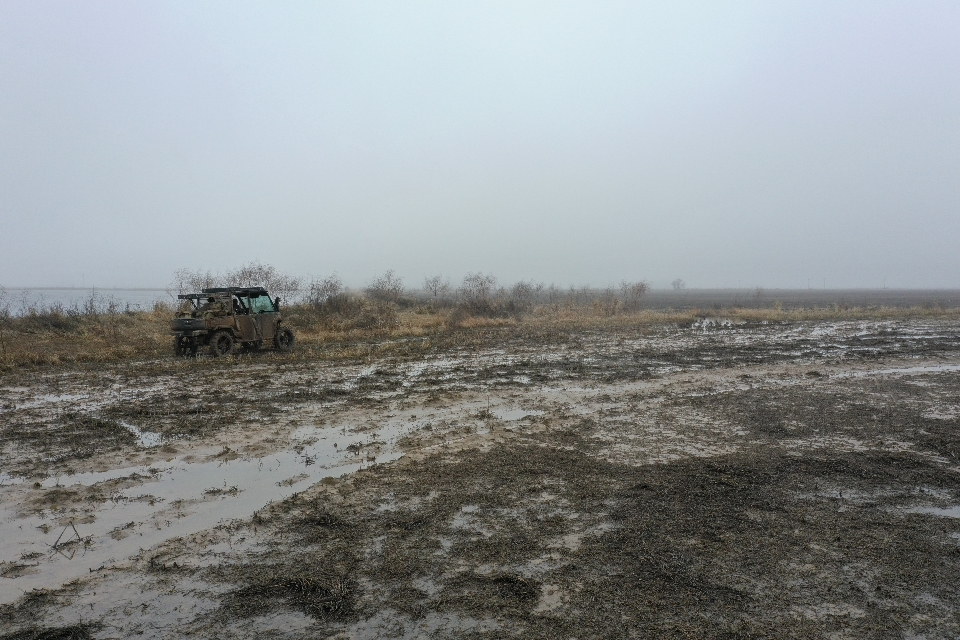 A muddy, flooded field under a gray sky with an off-road vehicle parked on the left.