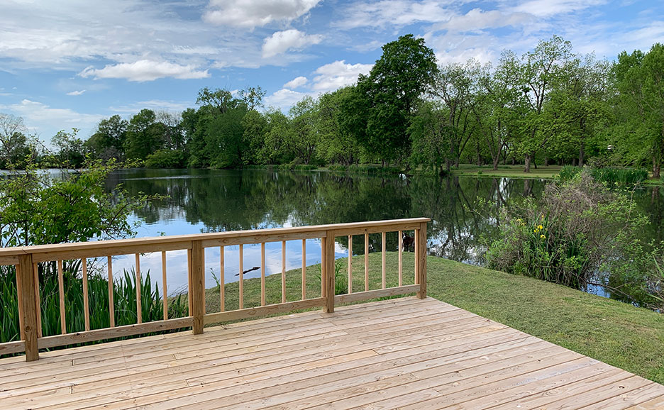 Wooden deck overlooking a calm pond with green trees and cloudy sky