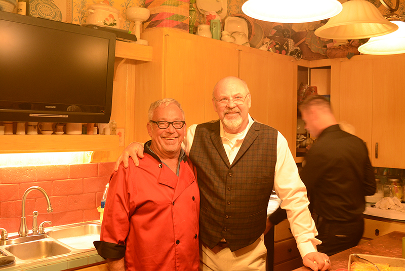 Two people smiling in a warm kitchen, standing behind a counter under hanging lights.