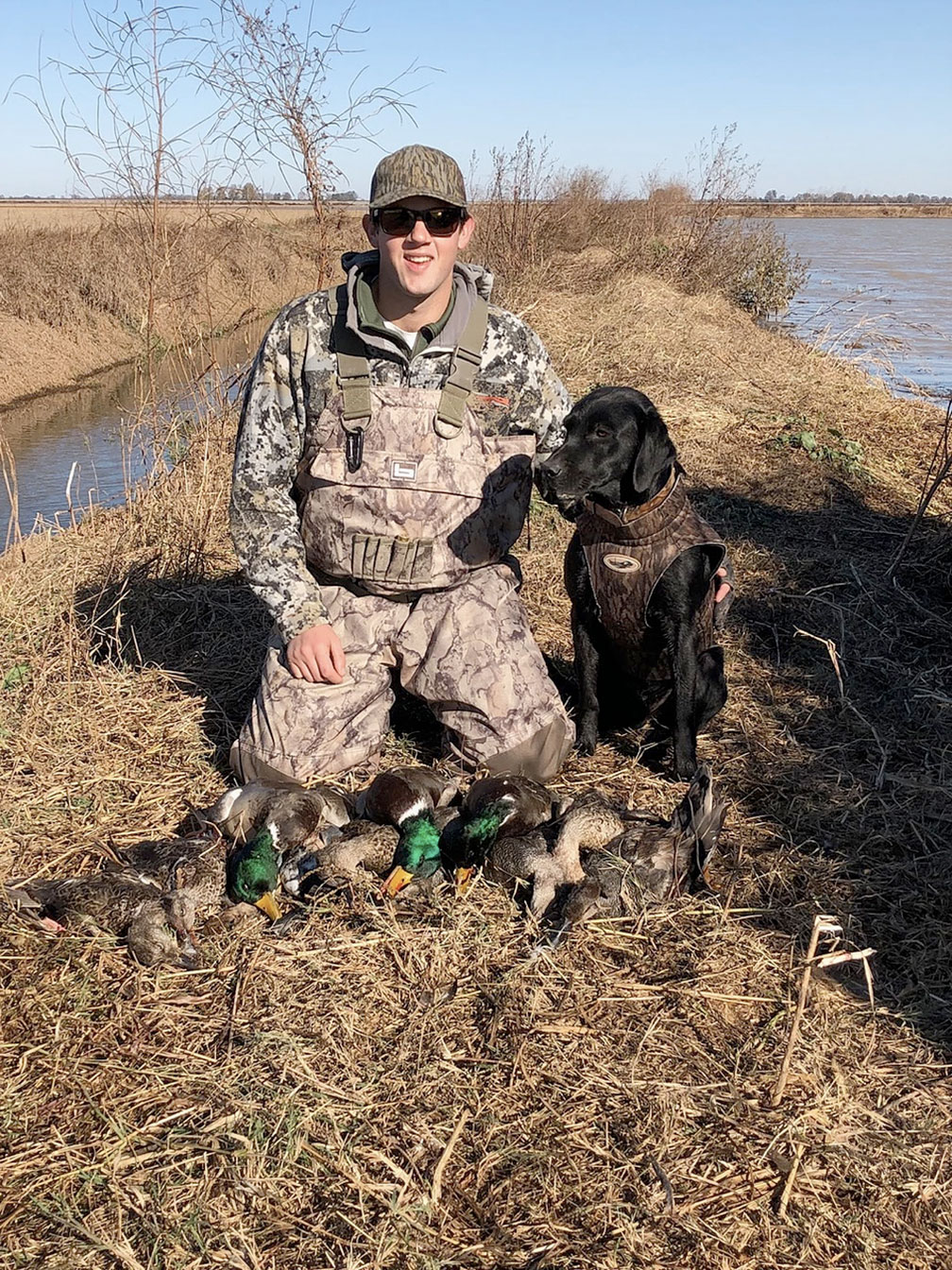Man in camouflage kneels beside a black dog in a grassy field near water under a clear sky.