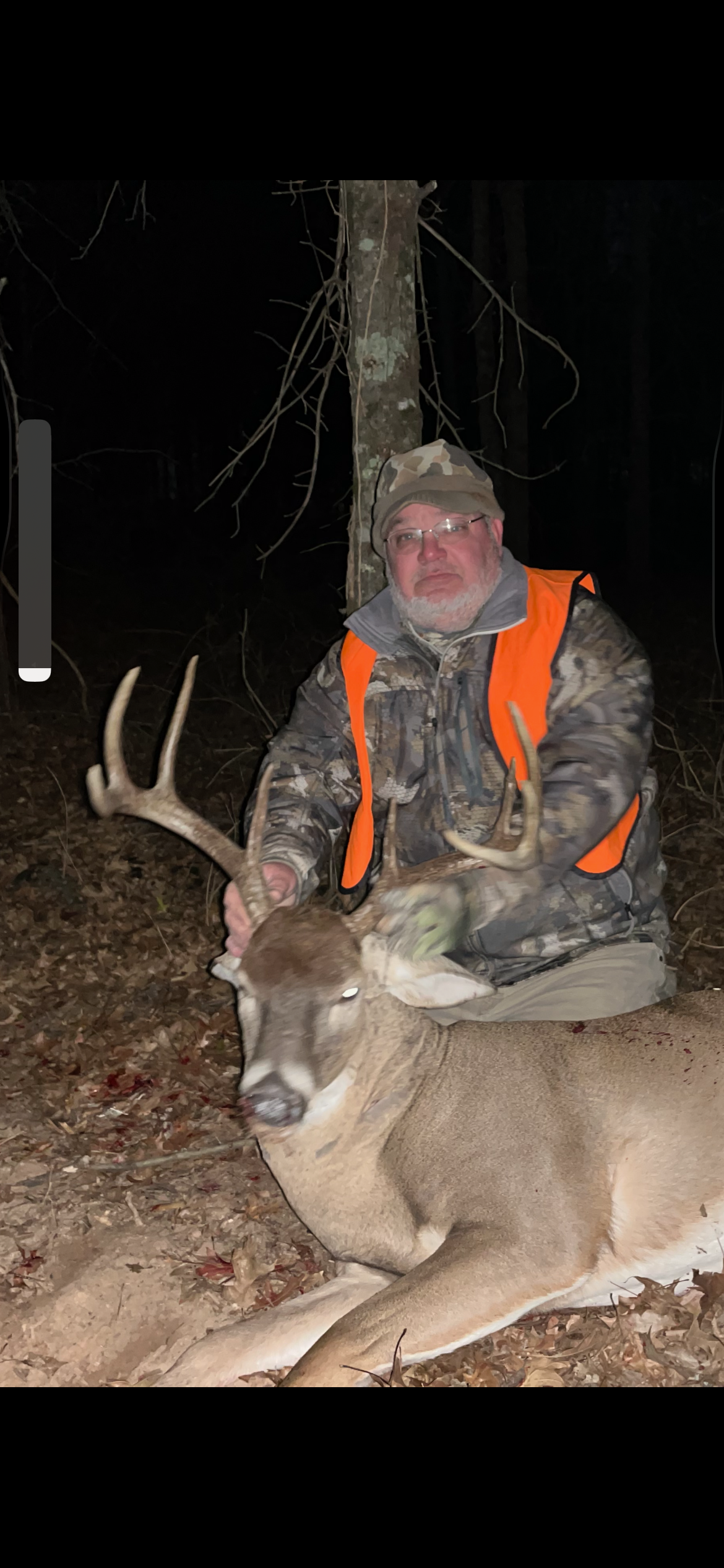 Hunter in orange vest kneels behind a large deer at night in the woods.