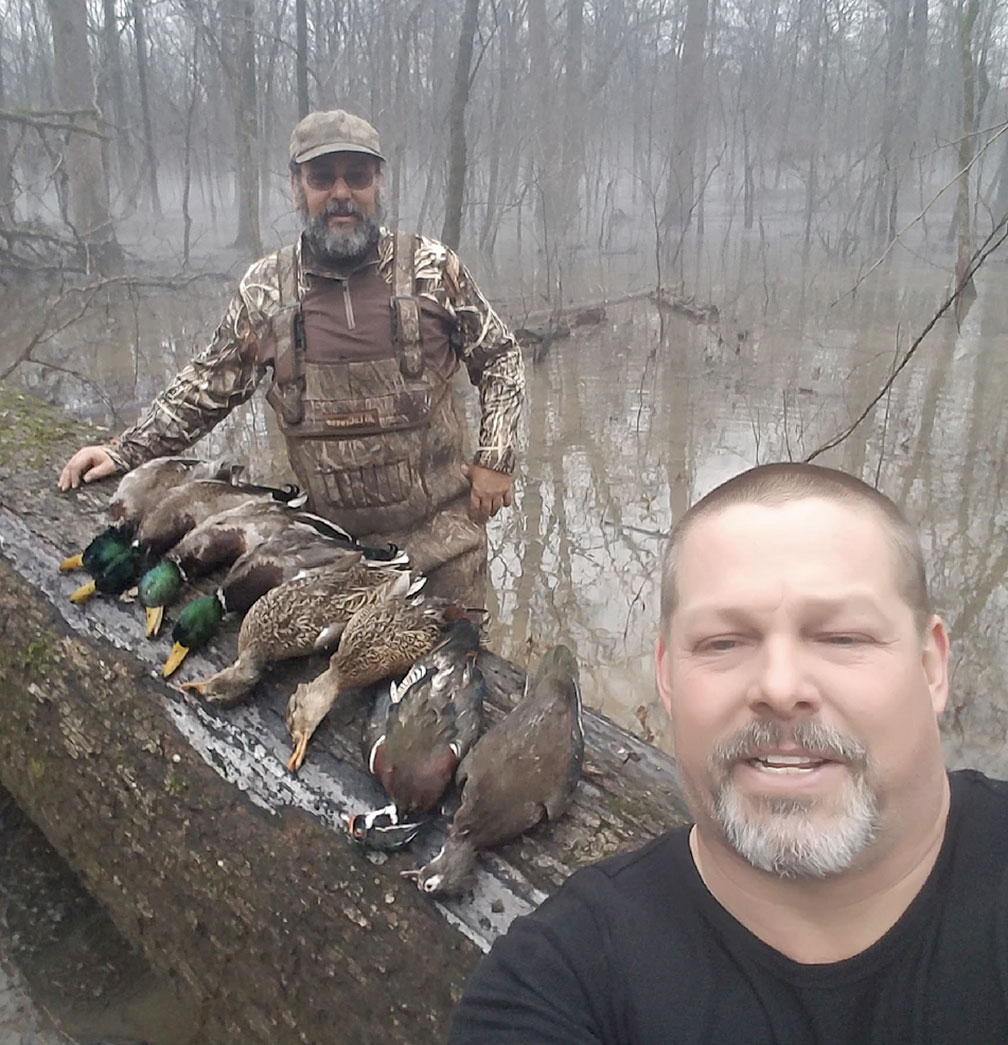 Two men pose by a log with four ducks in a wet, leafless woodland.