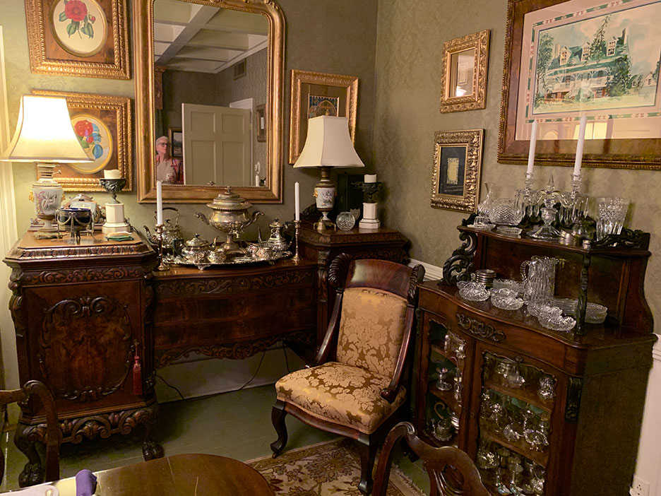 Elegant antique dining room with ornate wooden furniture, mirrors, lamps, and glassware displayed throughout
