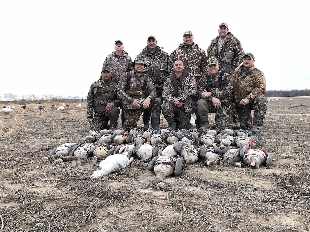 Group of camouflaged hunters posing behind a row of harvested ducks in a field.