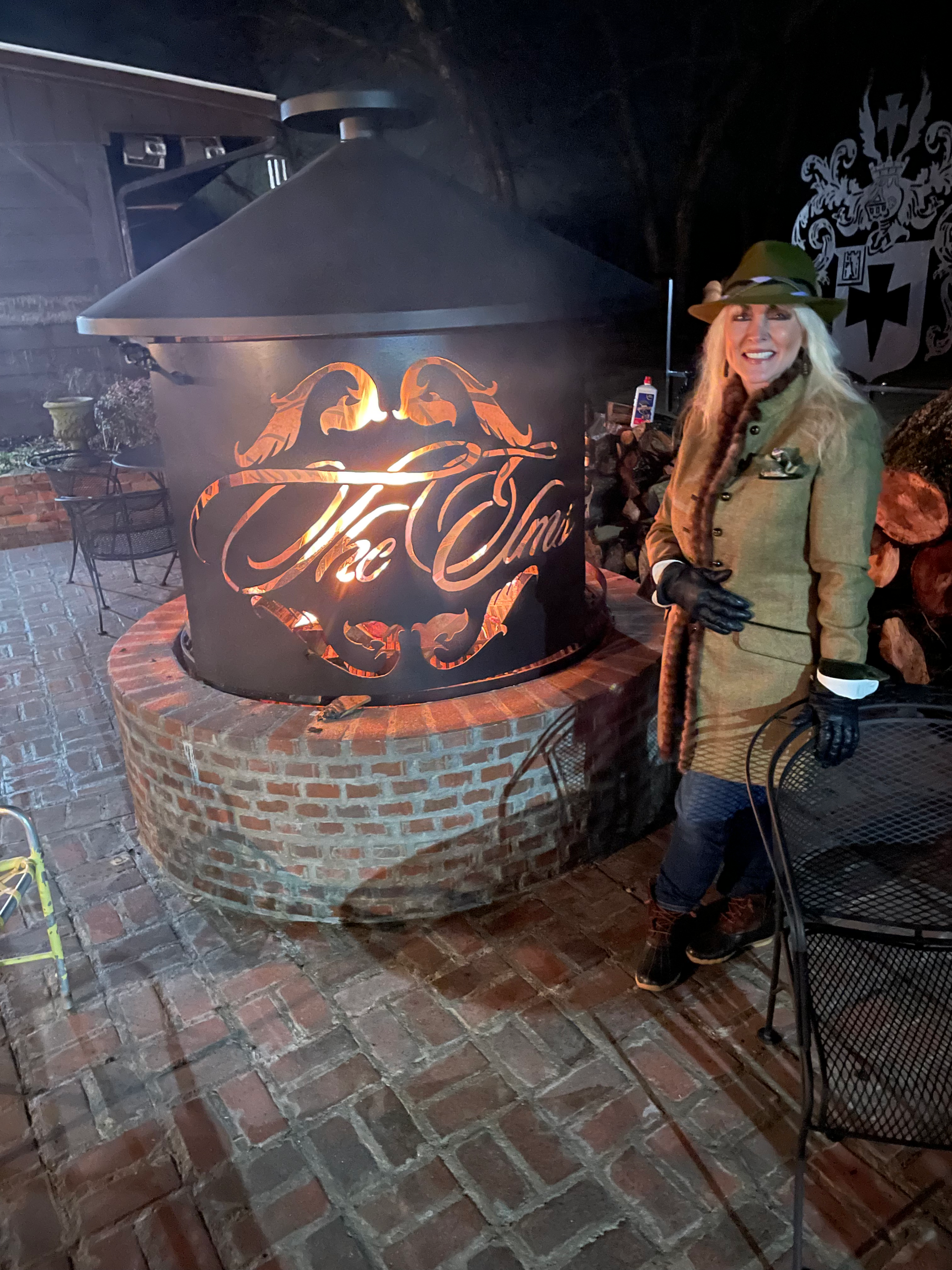 Woman in a green jacket stands beside a glowing fire pit with decorative script in a brick patio setting