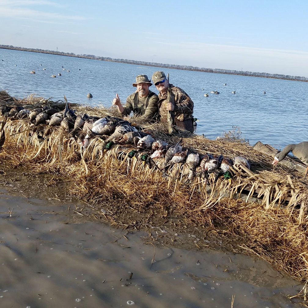 Two camouflaged hunters sitting behind a line of decoy geese beside a snowy shoreline and water