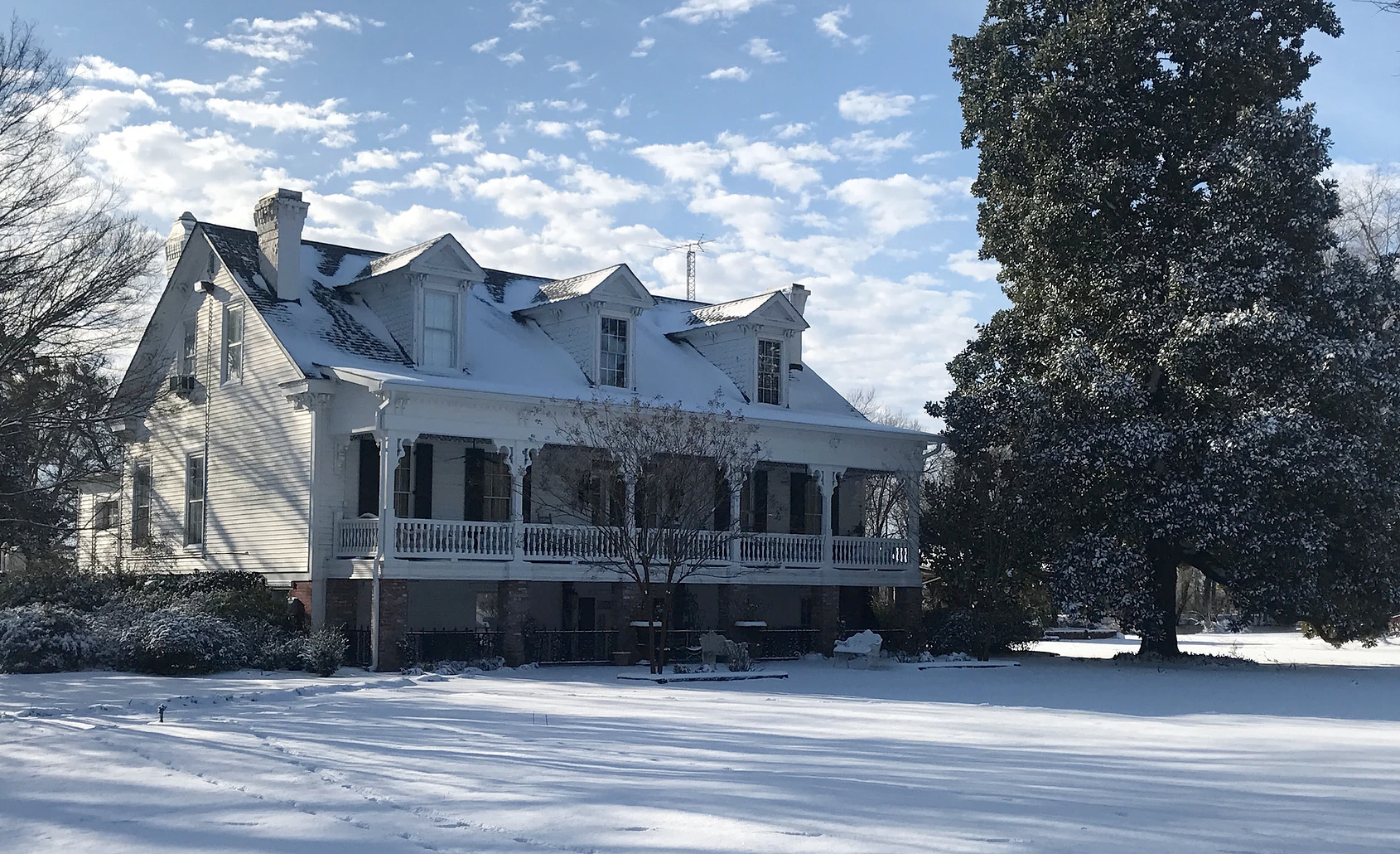 Snow-covered white house with a porch beside a large tree under a partly cloudy sky