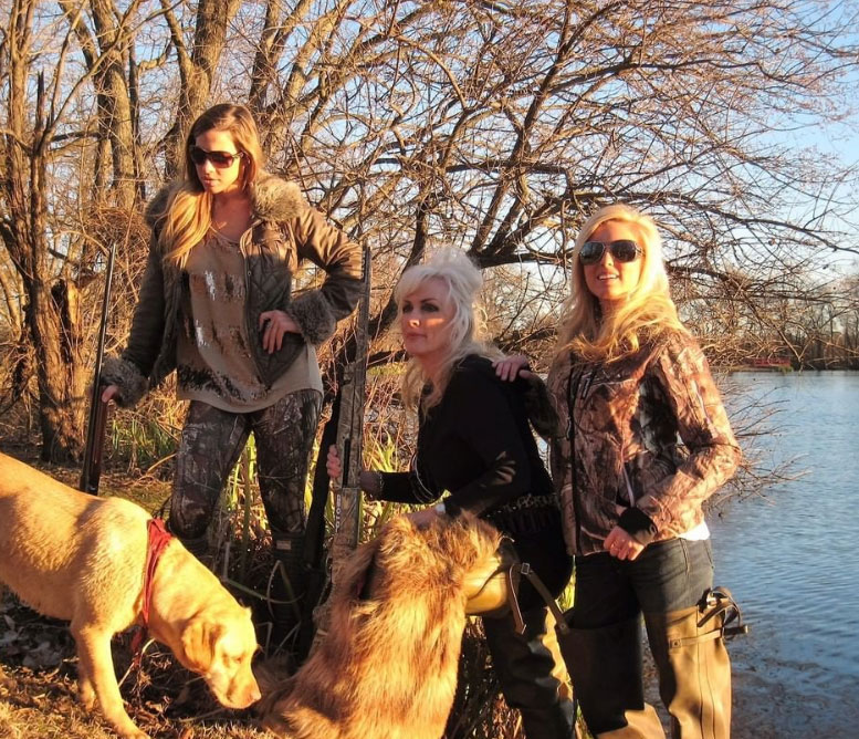 Three women with two dogs posing by a lake on a sunny day.