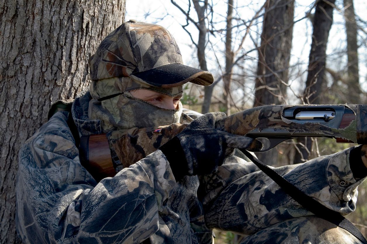 Camouflaged hunter aiming a shotgun from behind a tree in a winter woods.