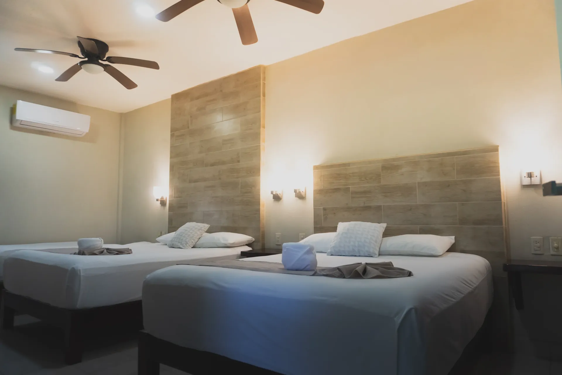 Habitación de hotel luminosa con dos camas bien hechas, iluminación cálida y ventilador de techo.