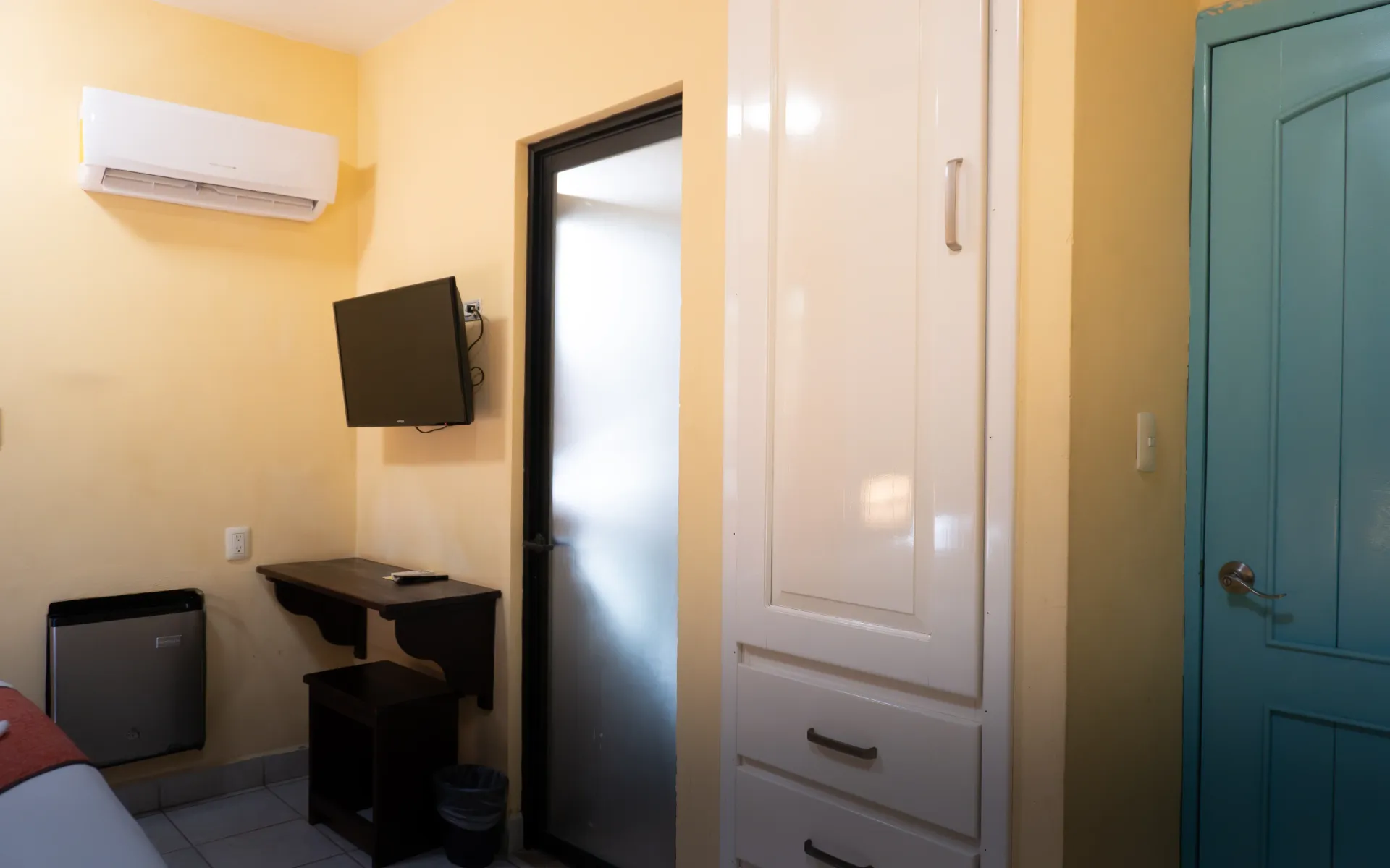 Habitación pequeña con televisor montado en la pared, escritorio, aire acondicionado y un armario blanco.