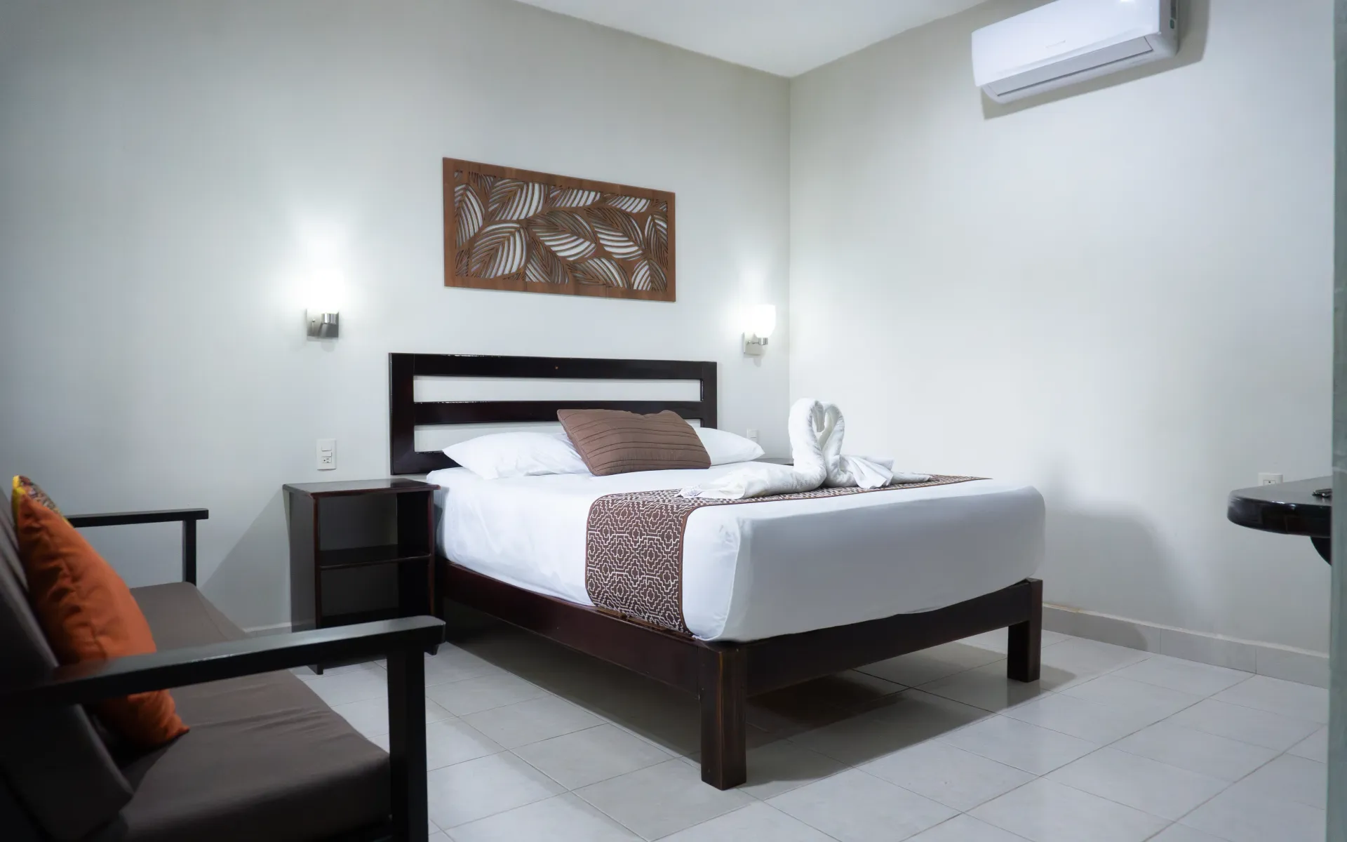 Habitación de hotel pequeña con cama, sofá, mesitas de noche, cuadros en la pared y aire acondicionado.