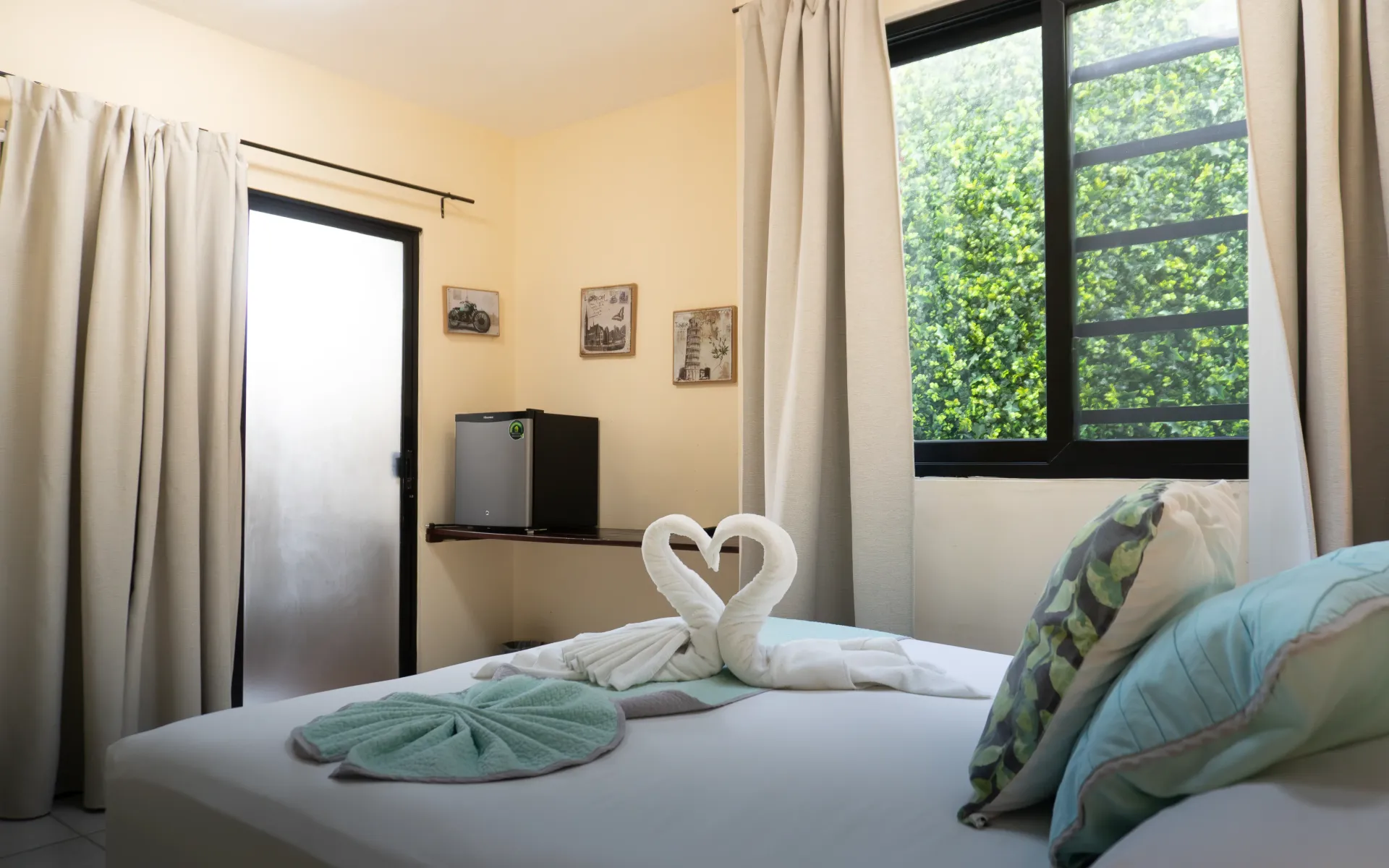 Habitación acogedora con cama blanca, cisnes de toalla, cortinas transparentes y una gran ventana con vegetación.