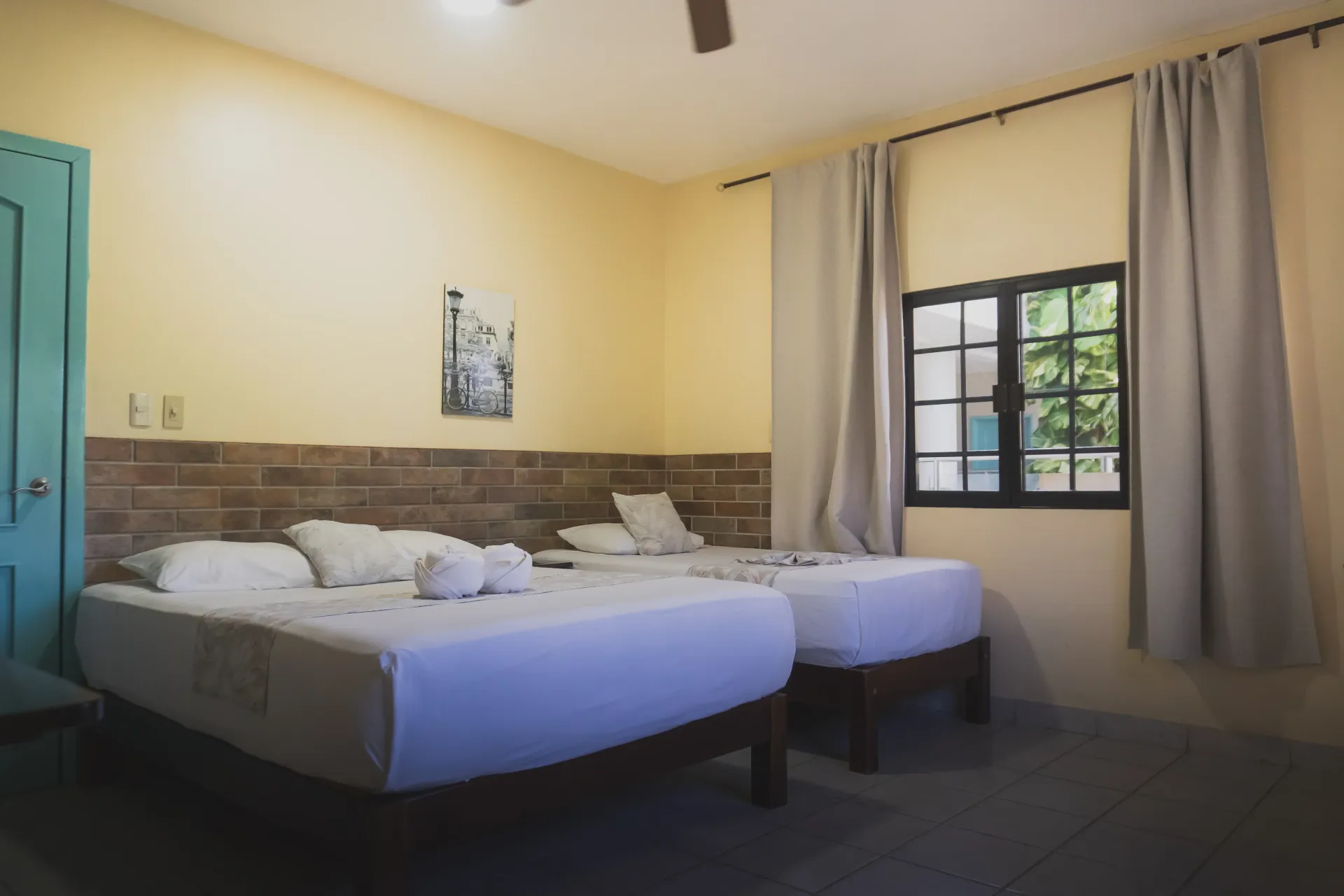 Habitación de hotel pequeña con dos camas individuales, paredes color crema y una ventana con cortinas.
