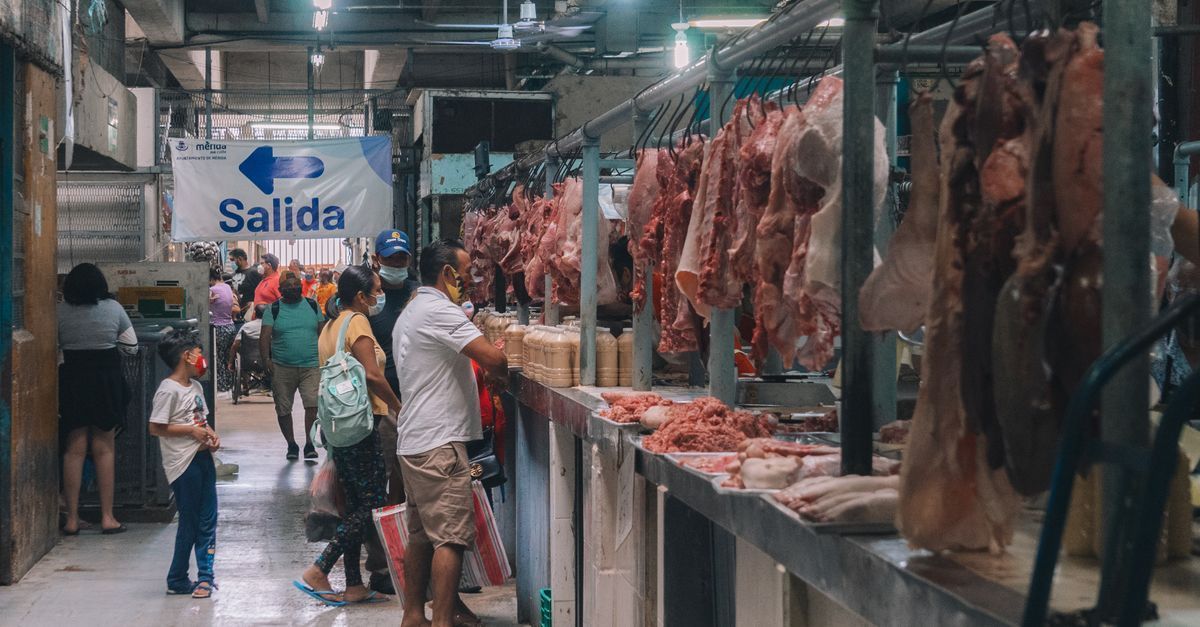 Pasillo de mercado cubierto con carne colgada, compradores y un letrero azul de 