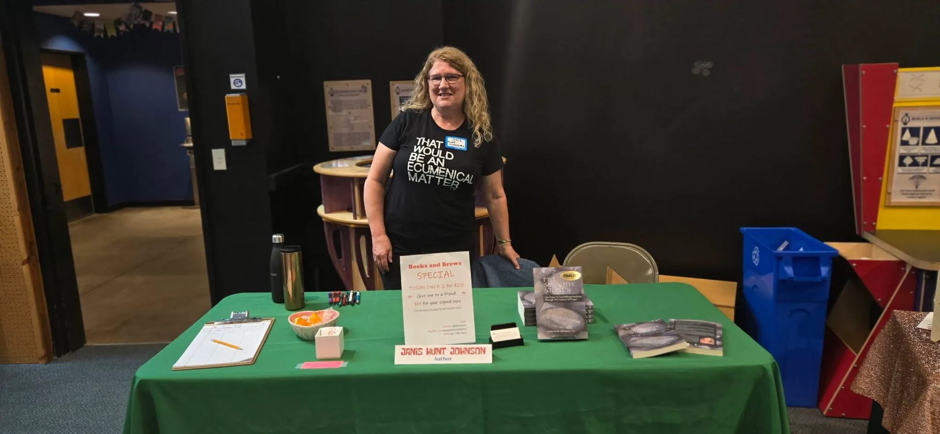 Woman behind a green table at an indoor event booth, with signs, brochures, and display items.