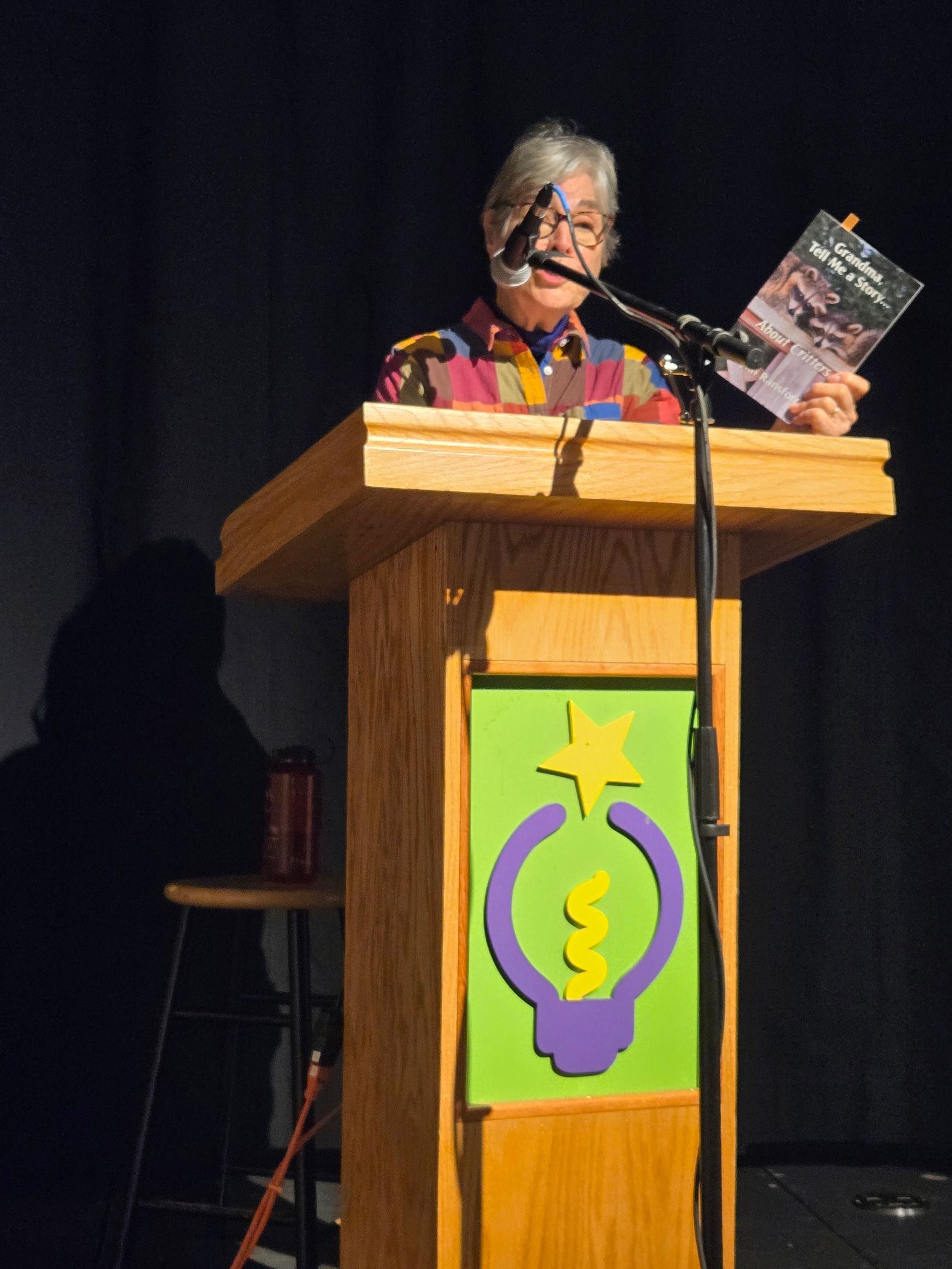 Speaker at podium holding a book on a dark stage, with a colorful sign on the lectern.