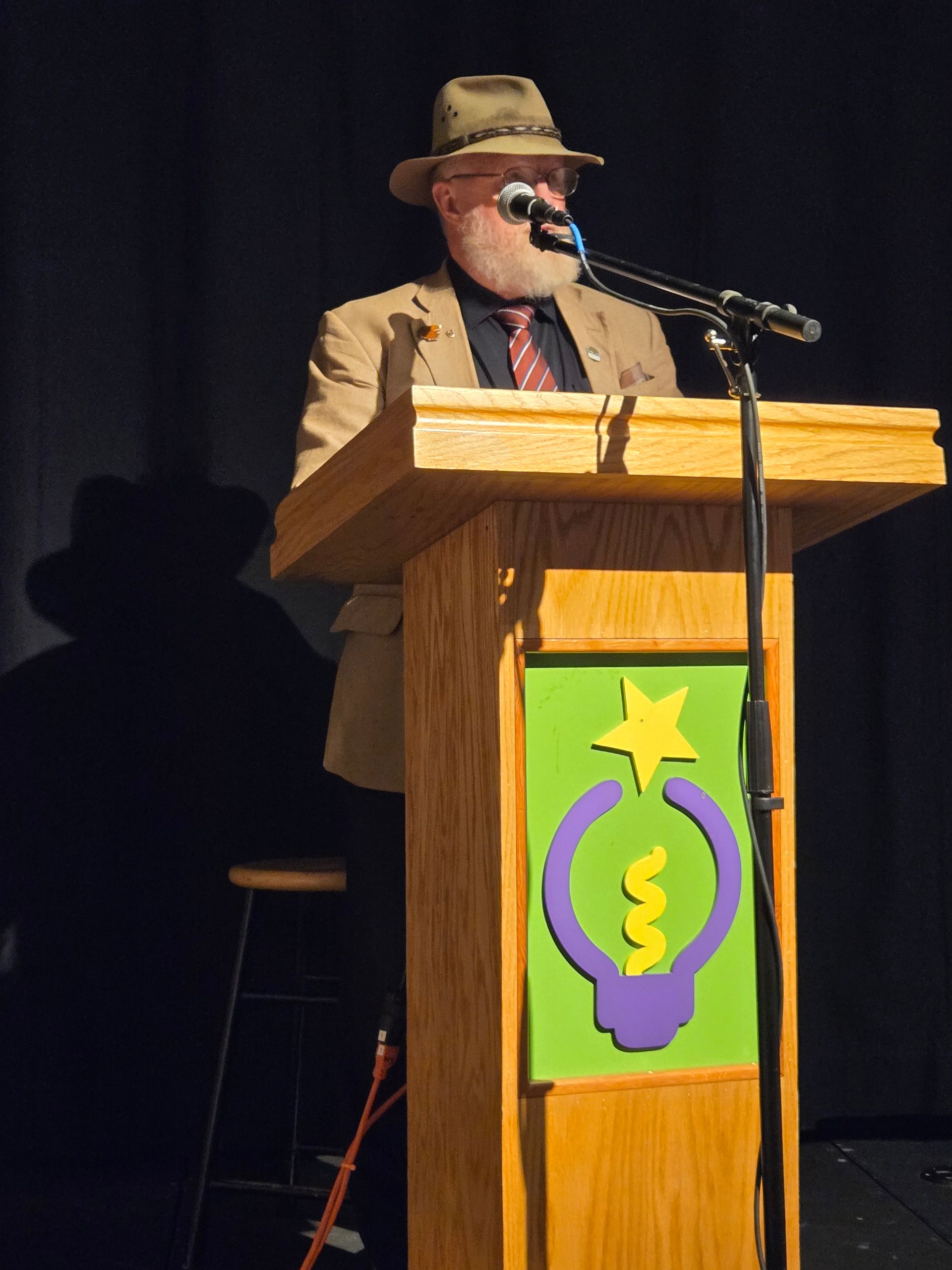 Man speaking at a wooden podium onstage, wearing a hat and glasses, with a green-and-purple lightbulb banner on the front.