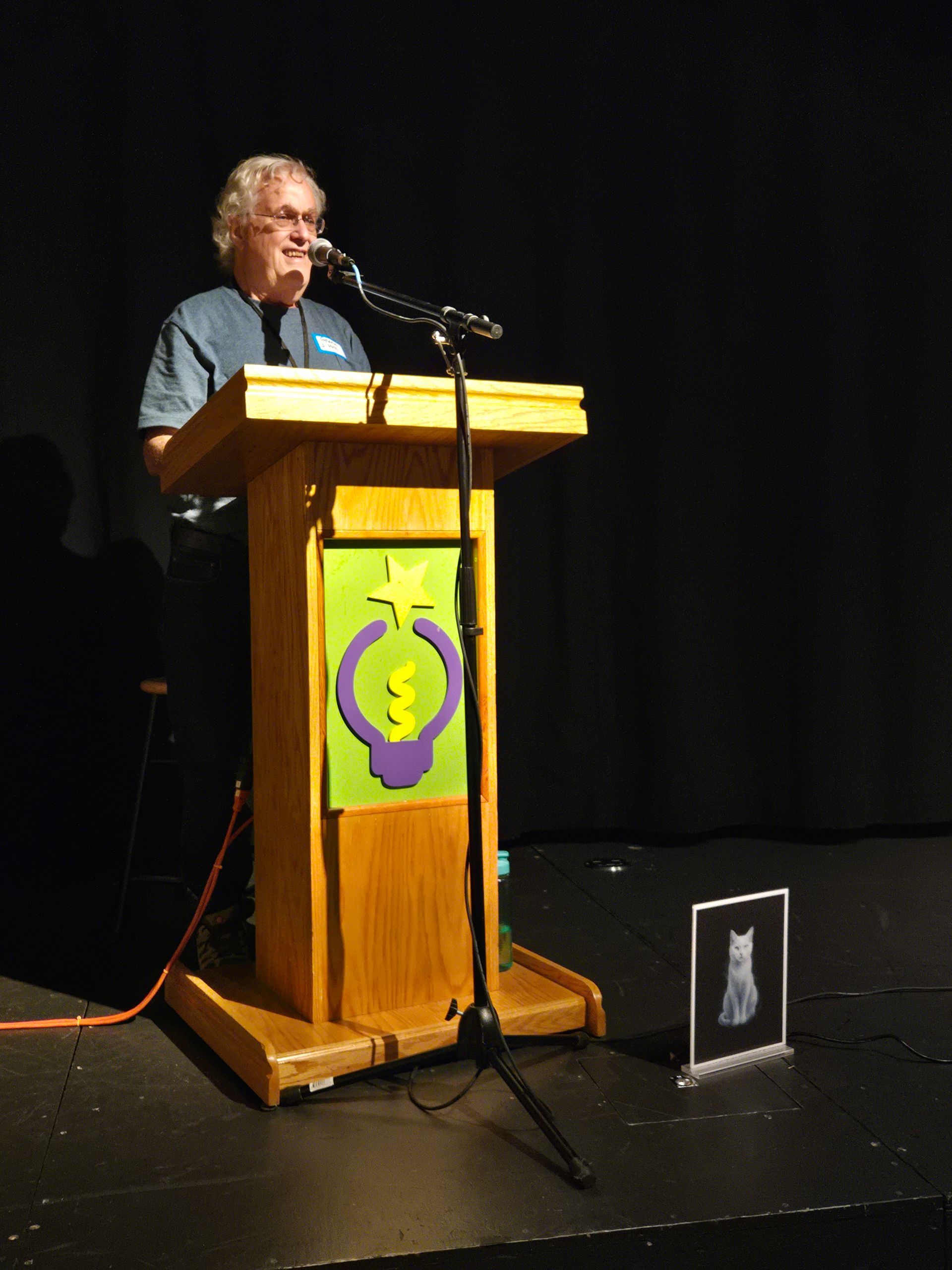 Person speaking at a wooden podium on a dark stage, with a colorful emblem on the front.