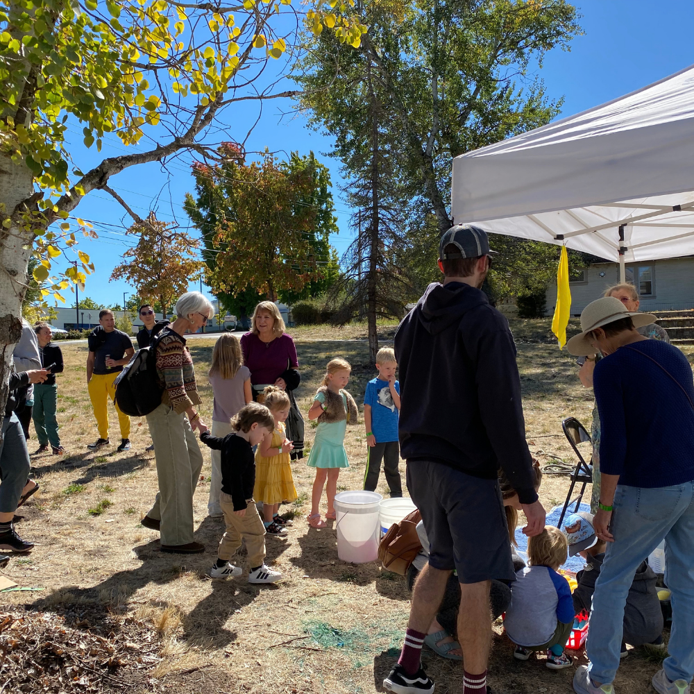 People gathered outdoors under trees near a white canopy tent on a sunny day.