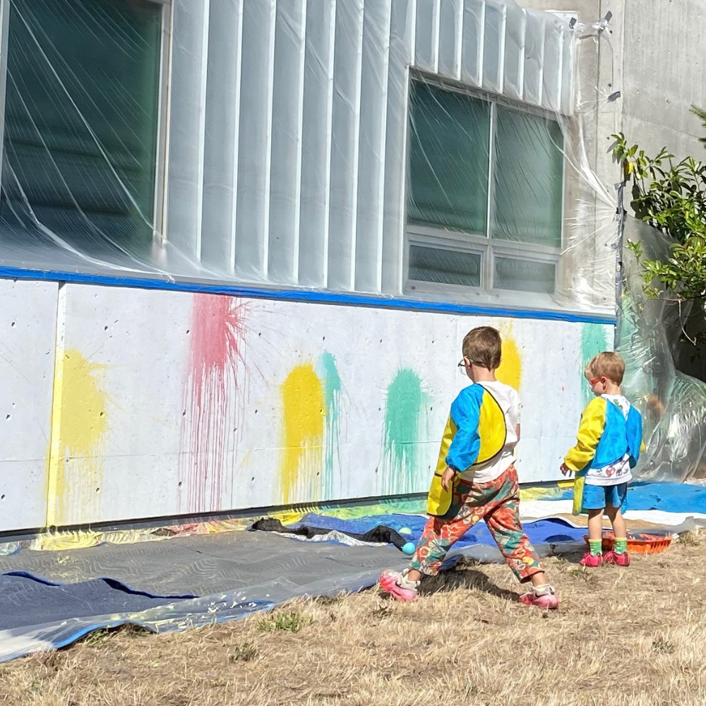 Two children paint colorful splotches on an outdoor wall