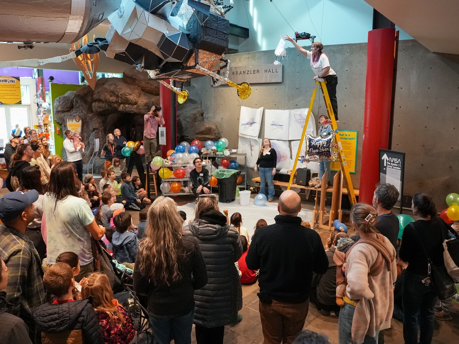 Crowd watches a person on a ladder throwing releasing egg drop challege crafts in an indoor space  at ScienceWorks