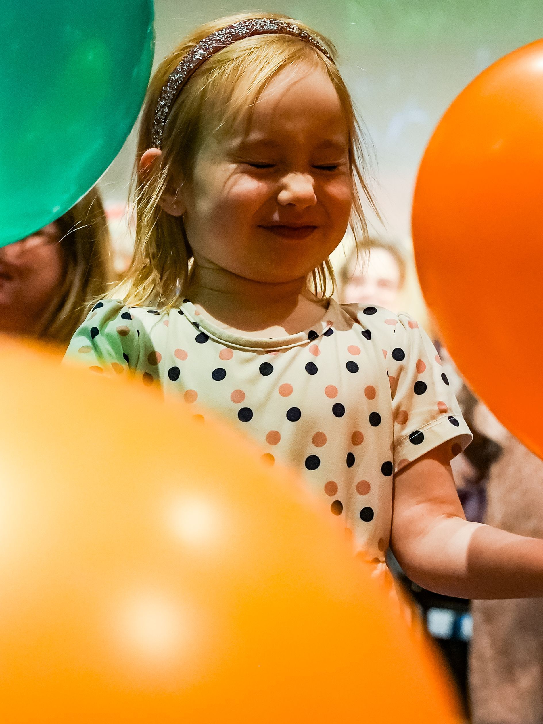 Girl with closed eyes smiles, surrounded by orange and green balloons at ScienceWorks.