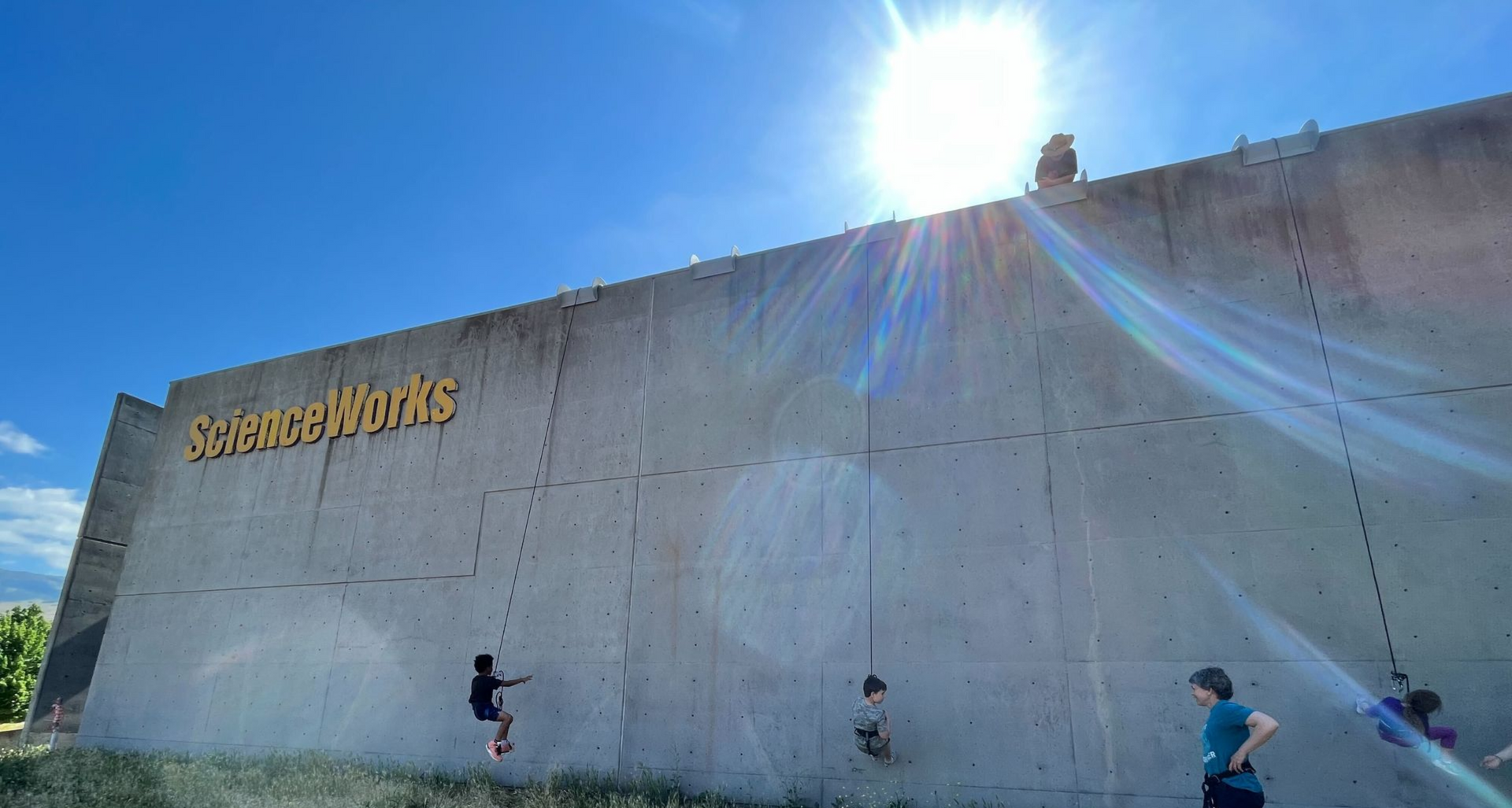 People rock climbing on a tall, outdoor wall labeled 