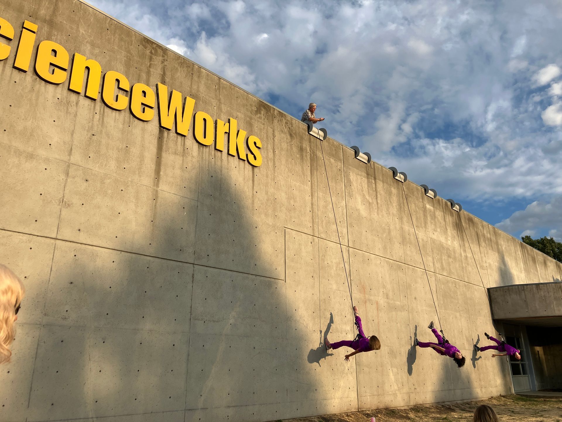 People in purple jumpsuits rappel down the exterior concrete wall of a building labeled 