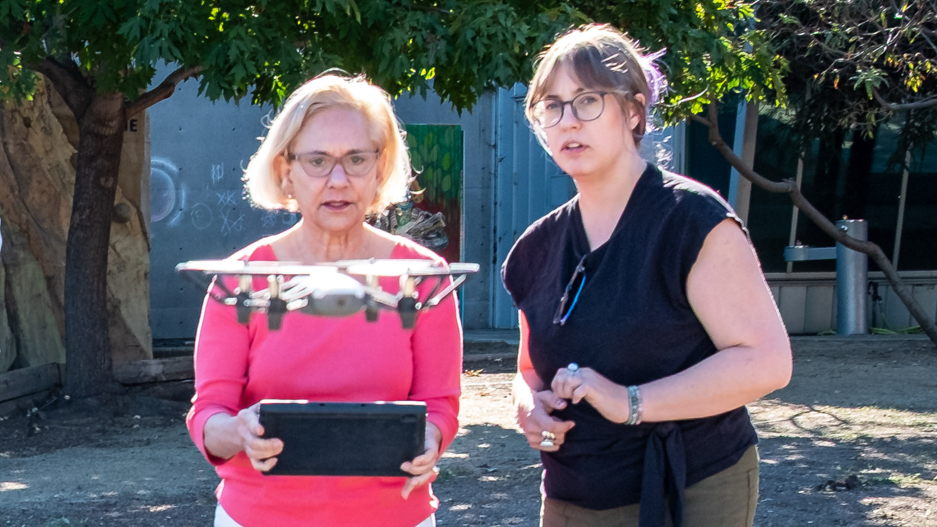 Two people outdoors flying a drone, one holding a tablet, with trees and a building behind them.