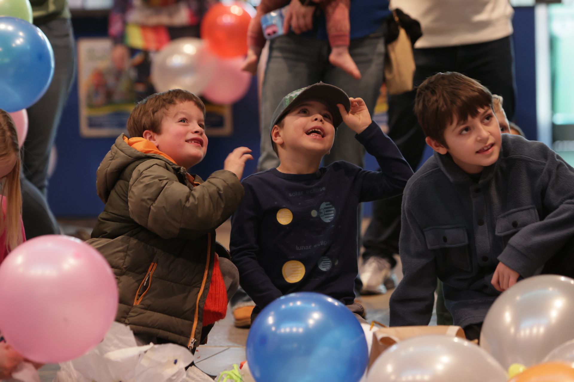Three boys looking up, surrounded by balloons; one adjusting hat, excited expressions. Indoors  at ScienceWorks.