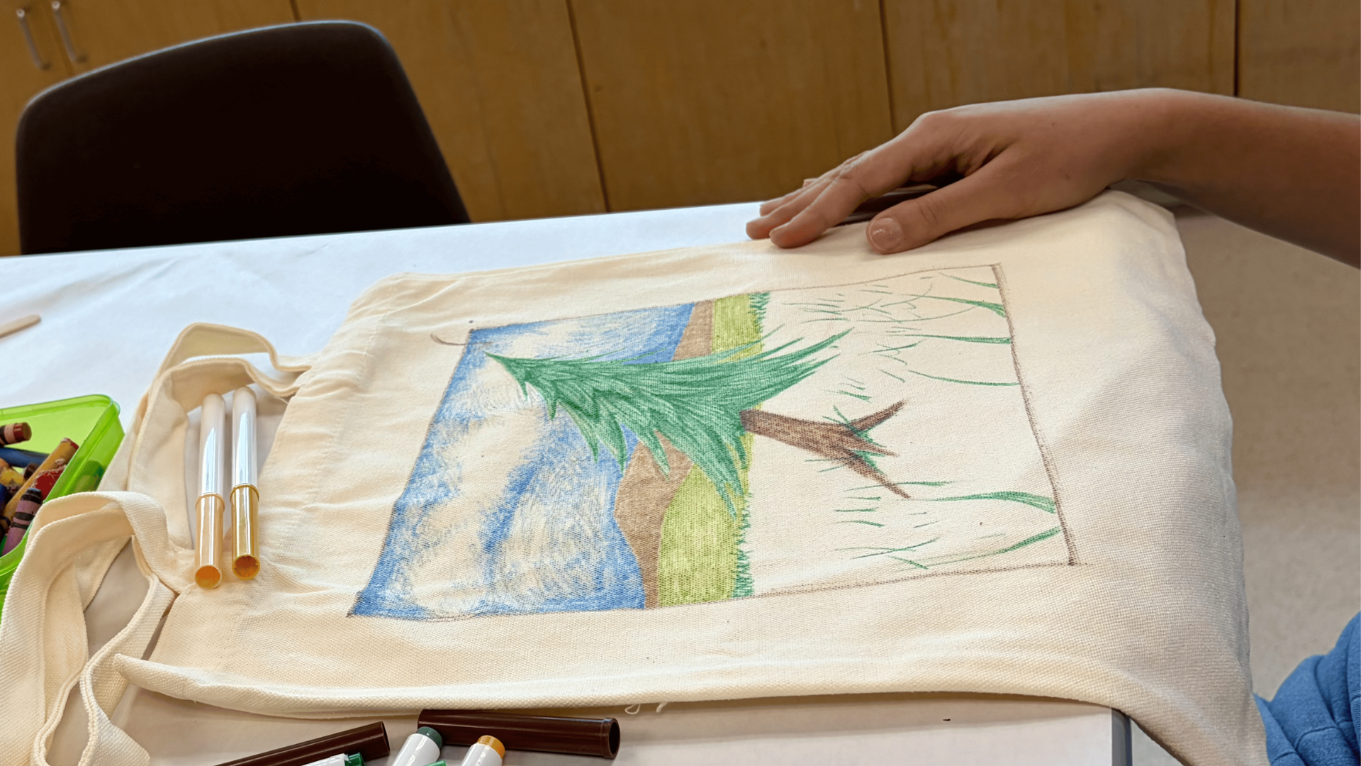 A visitor draws a landscape scene featuring a conifer on a cotton tote bage at Sparking Action