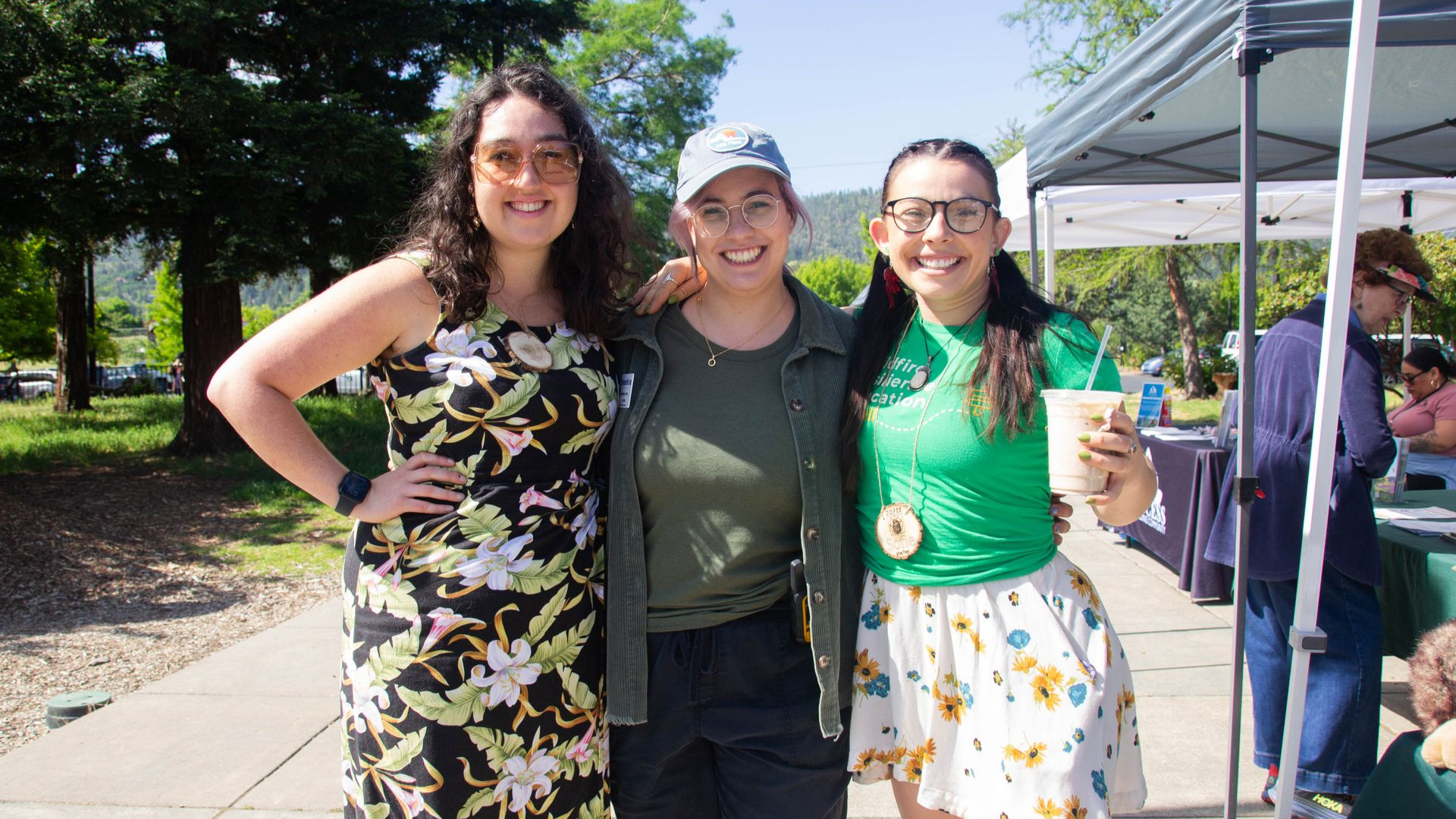 Three smiling people pose outdoors; one in a floral dress, a second in a hat, and the third holding a drink.