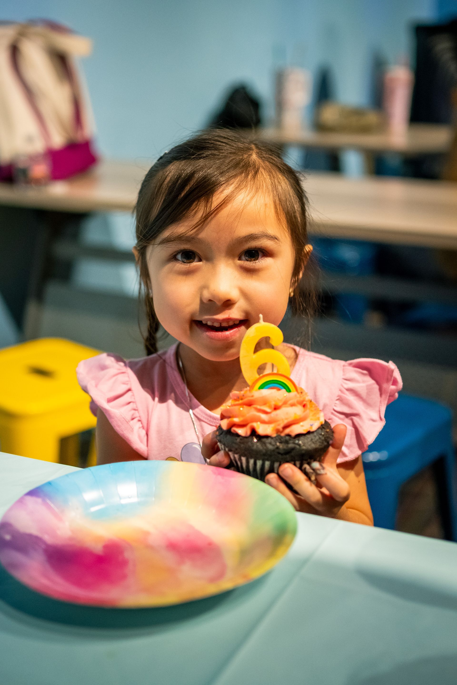 Child smiling at a table, holding a cupcake with a yellow number 6 candle and a colorful plate in front.