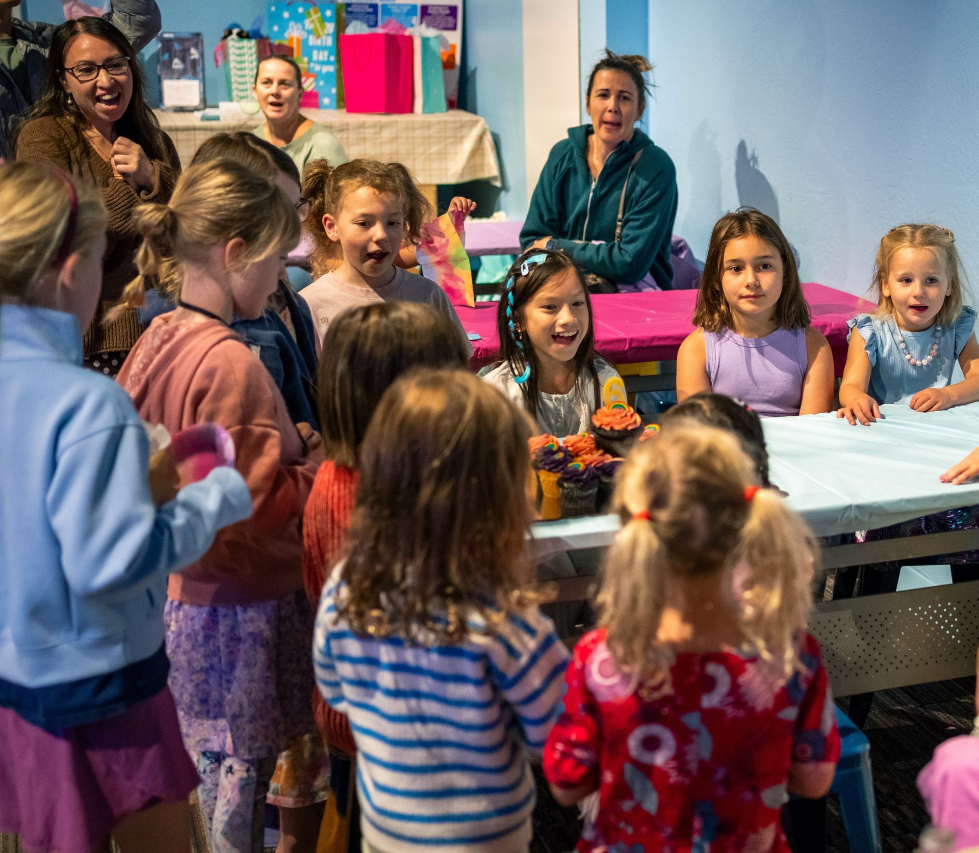 Children gathered around a table in a classroom, watching a demonstration with adults in the background.