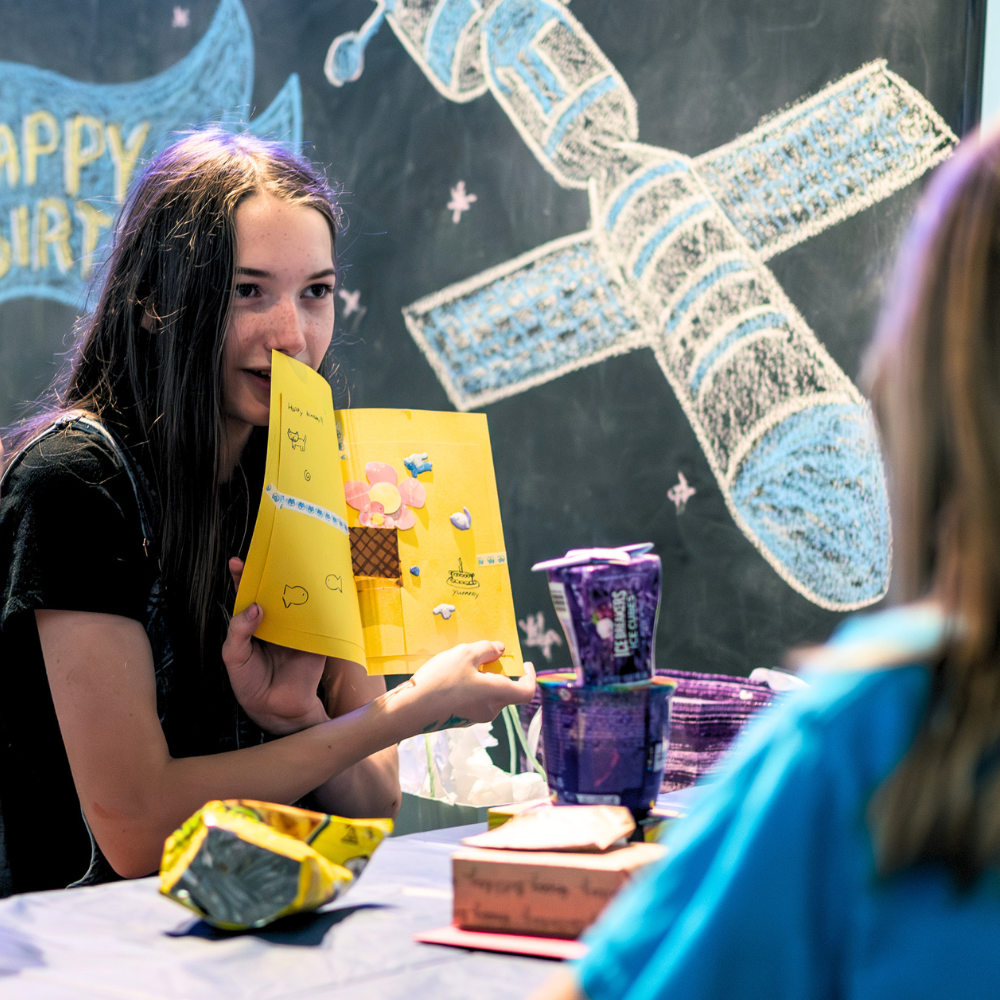Girl at birthday party holding a yellow card, with gift bags and chalk spaceship drawings on a blackboard backdrop
